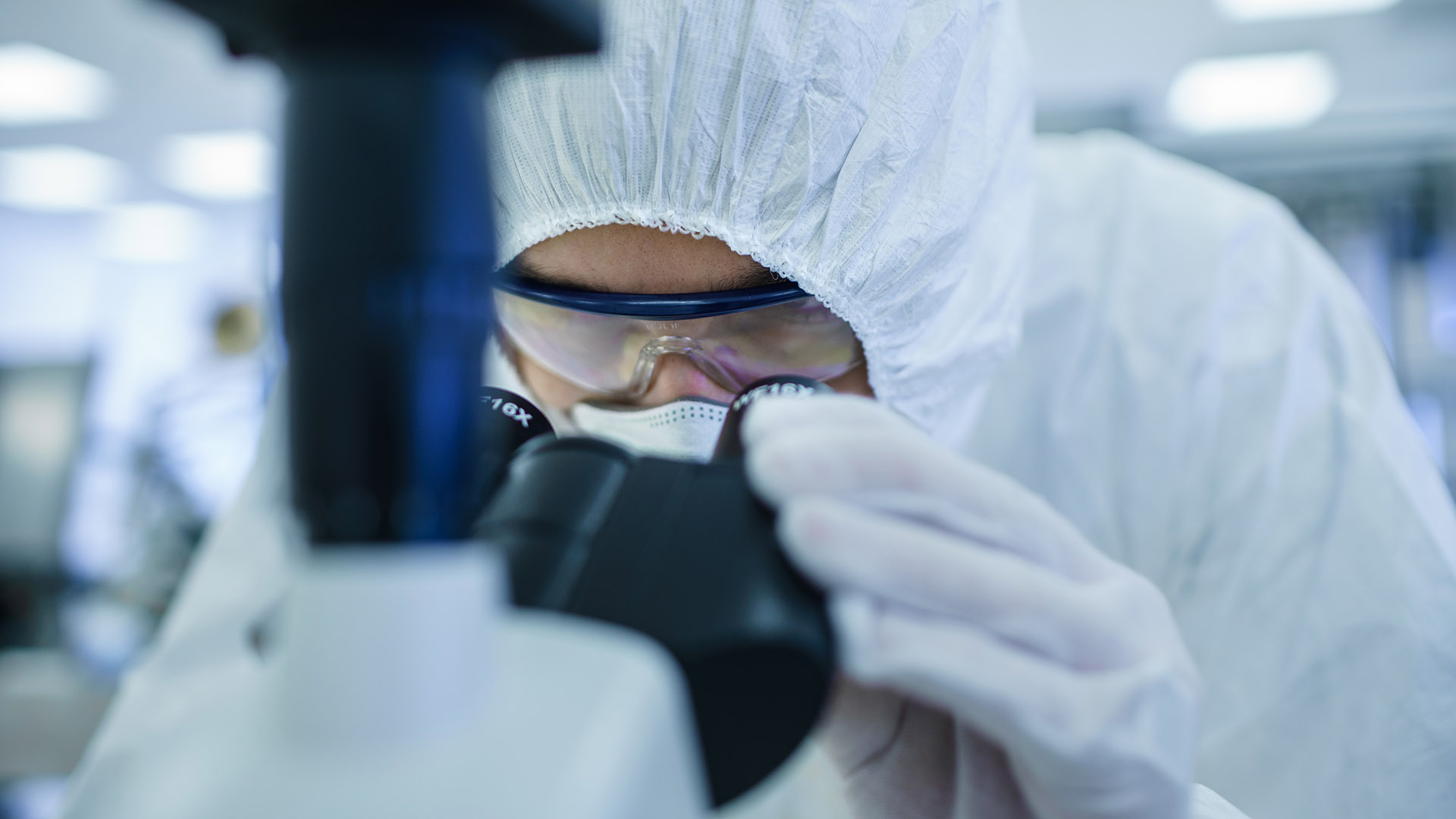 Close-up Shot of a Scientist in Protective Clothes Doing Research Uses Microscope and Personal Computer In Laboratory. Modern Manufactory Producing Semiconductors and Pharmaceutical Items.